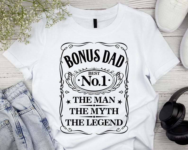 Pode incluir: Camiseta branca com um gr&aacute;fico preto que diz "BONUS DAD BEST No.1 THE MAN THE MYTH THE LEGEND". O design &eacute; emoldurado com linhas decorativas e estrelas. A camiseta &eacute; exibida em uma superf&iacute;cie branca com um par de jeans azuis, t&ecirc;nis brancos e fones de ouvido pretos.