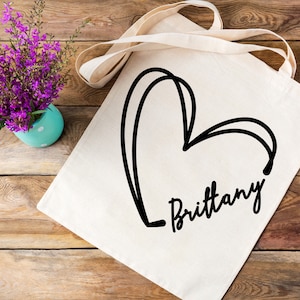 May include: A white canvas tote bag with a black hand-drawn heart and the name "Brittany" written underneath.