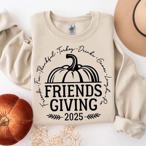 May include: A cream Friendsgiving sweatshirt featuring a black pumpkin design and the text "Friends Giving 2025". The design includes the words "Thankful, Turkey, Drinks, Games, Laughs, Pie, Fun" around the pumpkin.