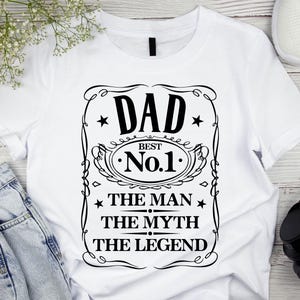 Pode incluir: Camiseta branca com um gr&aacute;fico preto que diz "DAD BEST No.1 THE MAN THE MYTH THE LEGEND" em uma moldura decorativa. A camiseta &eacute; exibida com jeans azuis, t&ecirc;nis brancos e fones de ouvido pretos.