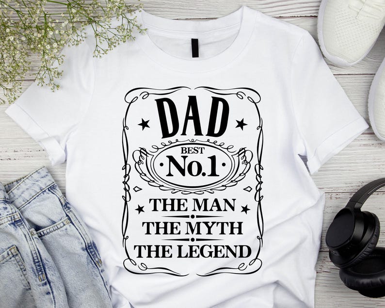 Pode incluir: Camiseta branca com um gr&aacute;fico preto que diz "DAD BEST No.1 THE MAN THE MYTH THE LEGEND" em uma moldura decorativa. A camiseta &eacute; exibida com jeans azuis, t&ecirc;nis brancos e fones de ouvido pretos.