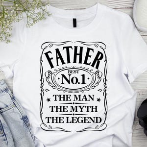 Pode incluir: Camiseta branca com um gr&aacute;fico preto que diz "FATHER BEST No.1 THE MAN THE MYTH THE LEGEND". O design &eacute; emoldurado com linhas decorativas e estrelas. A camiseta &eacute; exibida com jeans azuis, t&ecirc;nis brancos e fones de ouvido pretos.