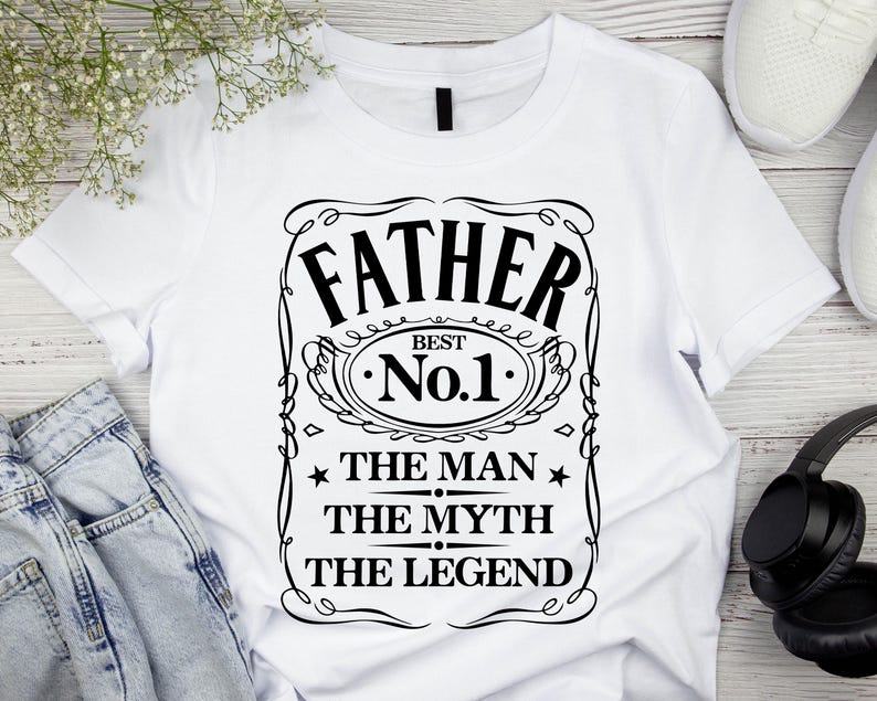 Pode incluir: Camiseta branca com um gr&aacute;fico preto que diz "FATHER BEST No.1 THE MAN THE MYTH THE LEGEND". O design &eacute; emoldurado com linhas decorativas e estrelas. A camiseta &eacute; exibida com jeans azuis, t&ecirc;nis brancos e fones de ouvido pretos.