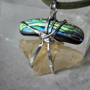 Puede incluir: Colgante de concha de abulón con colores iridiscentes, sujeto con alambre de plata y un cristal de cuarzo transparente. El colgante cuelga de un cordón verde.