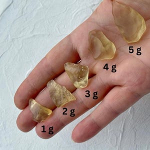 Puede incluir: Cinco piedras translúcidas de color ámbar, de forma irregular, se muestran en una mano. Cada piedra está etiquetada con su peso en gramos: 1 g, 2 g, 3 g, 4 g y 5 g. Las piedras varían en tamaño y forma.
