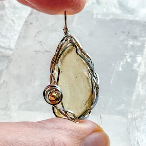 Puede incluir: Un colgante artesanal con una piedra en forma de lágrima de color ámbar claro, encerrada en un marco de alambre plateado. Una pequeña cuenta dorada está incorporada en el diseño. El colgante está suspendido de un lazo de alambre plateado.