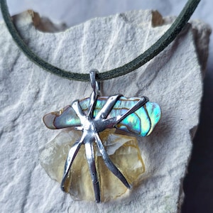 Puede incluir: Collar artesanal con un colgante de concha de abulón pulida con colores iridiscentes azul, verde y dorado, engarzado en metal plateado. El colgante está suspendido de un cordón verde oscuro.