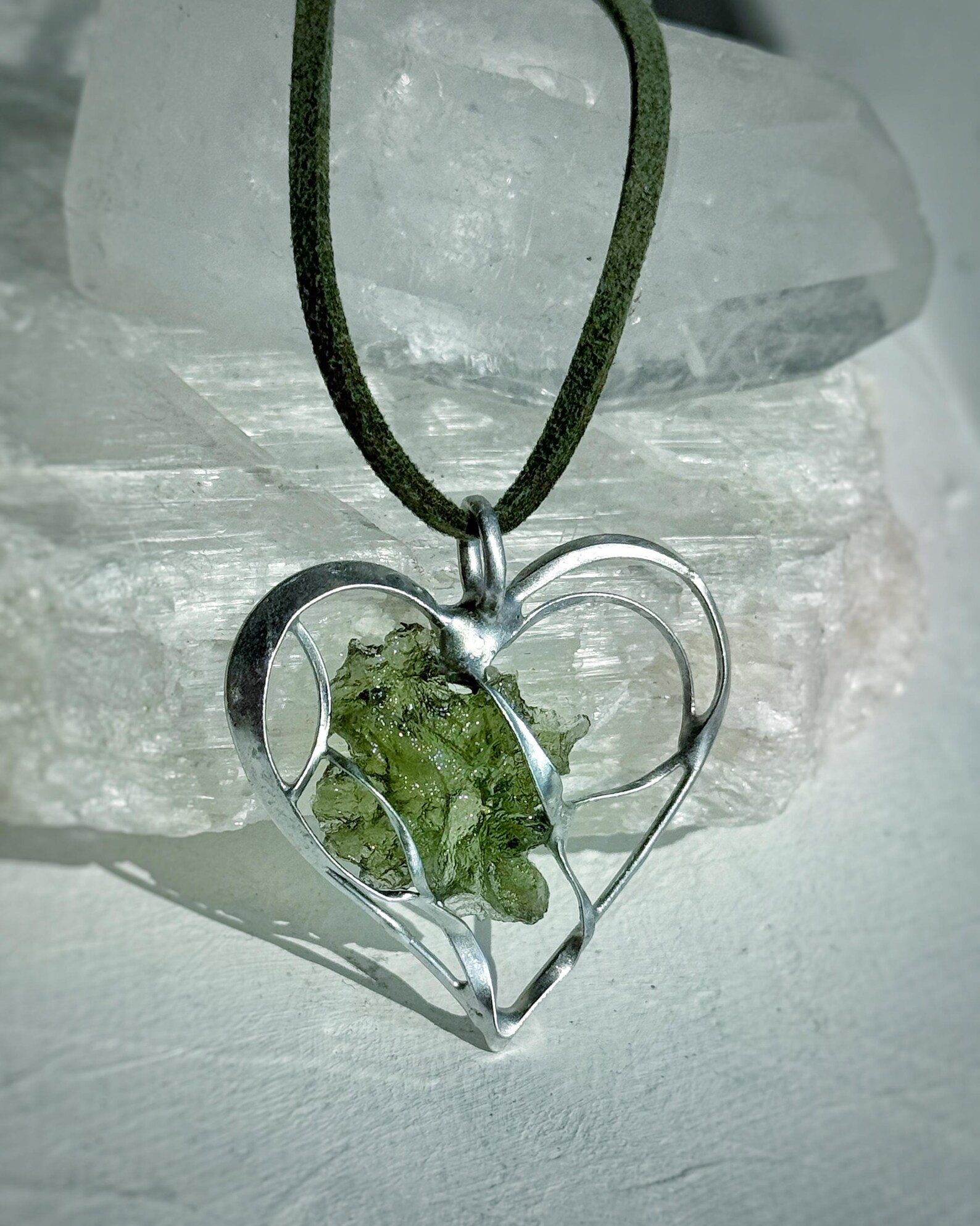 Raw moldavite heart pendant on green cord in silver setting, displayed against clear quartz background.