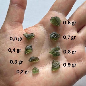 May include: A hand displays several pieces of green moldavite, each labeled with its weight in grams. The pieces vary in size and shape, ranging from 0.2 gr to 0.9 gr. The moldavite has a natural, rough texture.