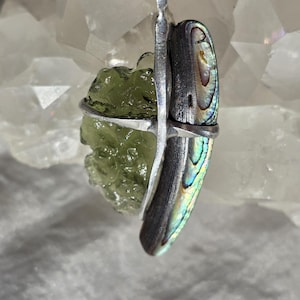 Puede incluir: Un collar con colgante que presenta una piedra rugosa de color verde claro y una concha de abulón iridiscente, ambas encapsuladas en un engaste de metal plateado. El collar cuelga de un cordón verde.