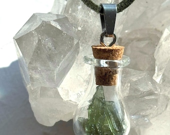 Op de afbeelding: Een kleine hanger van een doorzichtig glazen flesje met daarin een grillig stuk doorschijnende groene steen. Het flesje is afgesloten met een lichtbruine kurk en is voorzien van een zilverkleurig metalen oogje aan een donker olijfgroen koord.