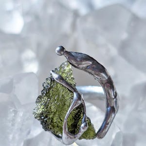 May include: A silver ring featuring a rough-cut, olive-green gemstone. The ring's band has an organic, flowing design that wraps around the stone. The gemstone is irregularly shaped, adding to the ring's unique aesthetic.