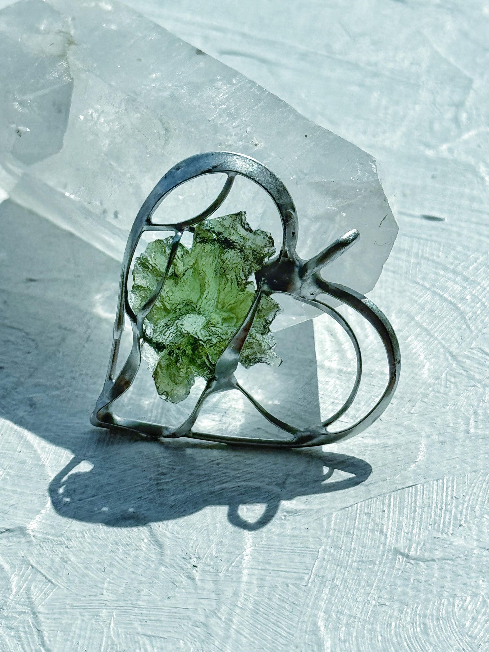 Raw moldavite heart pendant in silver setting with abstract design, displayed on a white crystal background.