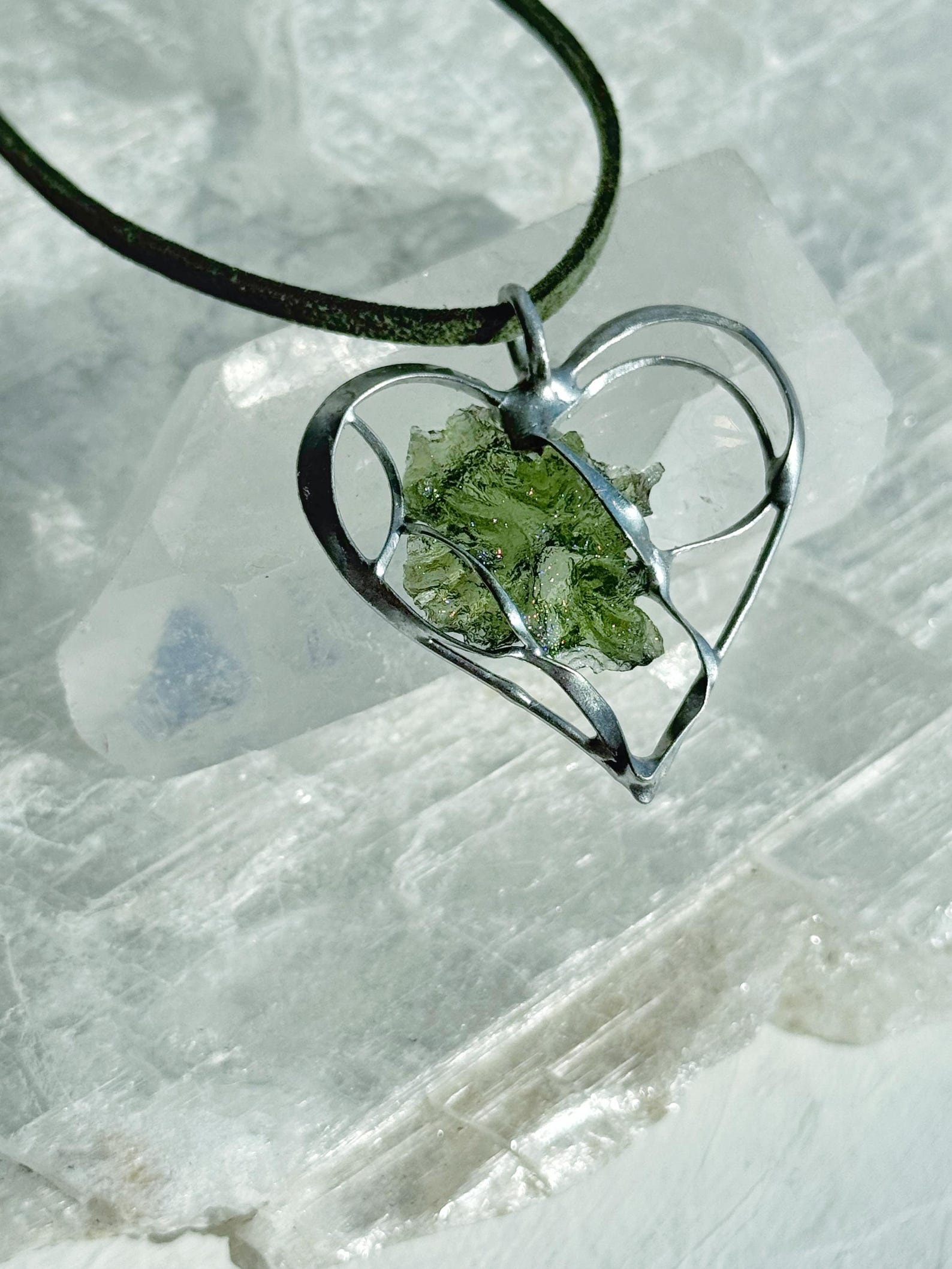 Raw moldavite heart pendant on green cord in silver setting, displayed against clear quartz background.