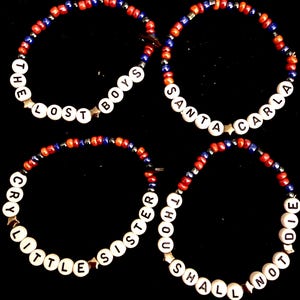 May include: Four beaded bracelets with white letter beads spelling out phrases like "THE LOST BOYS" and "SANTA CARLA". The bracelets feature red, blue, and silver beads. Each bracelet has a star-shaped bead.