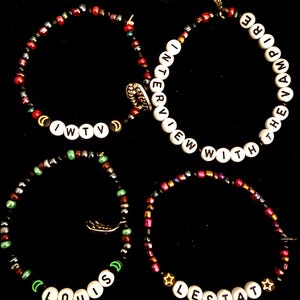 May include: Four beaded bracelets with white letter beads spelling out "IWTV", "INTERVIEW WITH THE VAMPIRE", "LOUIS", and "LESTAT". The bracelets feature various colored beads and charms, set against a black background.