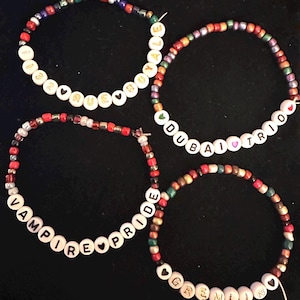 Könnte beinhalten: Vier farbige Perlenarmbänder mit weißen Perlen, die "TRUE YALE", "DUBAI I TRIO", "VAMPIRE PRIDE" und "GREMLIN" buchstabieren.