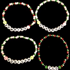 Puede incluir: Cuatro pulseras circulares de cuentas con pequeñas cuentas redondas en colores alternos rojo, verde y rosa claro. Tres pulseras incluyen cuentas de letras blancas que deletrean ZOMBIES, SEABROOK, ADDISON y ZED, acentuadas con pequeñas cuentas en forma de estrella y corazón.