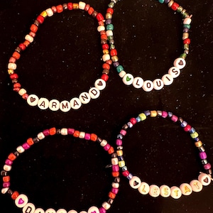 Könnte beinhalten: Vier bunte Perlenarmbänder mit weißen Buchstabenperlen, die die Namen "Armando", "Louis", "Daniel" und "Lestat" buchstabieren. Jedes Armband hat einen kleinen Herzanhänger.
