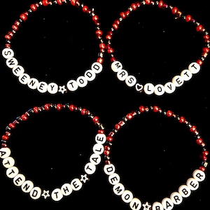 Könnte beinhalten: Vier Armbänder mit roten und schwarzen Perlen. Jedes Armband hat weiße Buchstabenperlen, die Sätze bilden: "SWEENEY TODD", "MRS LOVETT", "ATTEND THE TALE" und "DEMON BARBER". Sterne und ein Herz akzentuieren die Armbänder.
