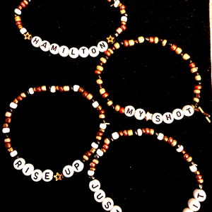 Peut inclure: Quatre bracelets de perles avec des lettres blanches épelant "HAMILTON", "MY SHOT", "RISE UP" et "JUST YOU WAIT". Les bracelets ont des perles marron, blanches et dorées, avec des accents en forme d'étoile. Le fond est noir.