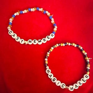 Puede incluir: Dos pulseras de cuentas con cuentas blancas y letras negras. La primera pulsera dice "CAST MEMBER" y la segunda pulsera dice "MAGIC MAKER".