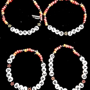 May include: Four beaded bracelets featuring red, orange, and yellow seed beads with white letter beads. The bracelets display the text SINNERS, CLUB JUKE, WE ARE ALL SINNERS, and DANCE WITH THE DEVIL, accented with small metallic star-shaped beads.