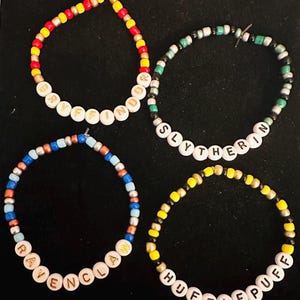 Puede incluir: Cuatro pulseras de cuentas con los nombres de las casas de Hogwarts: Gryffindor, Slytherin, Ravenclaw y Hufflepuff. Cada pulsera está hecha con cuentas de diferentes colores y presenta el nombre de la casa en letras doradas.