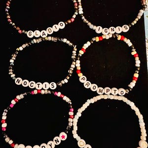 Puede incluir: Seis pulseras de cuentas de colores con diferentes nombres escritos en ellas. Los nombres son Gladio, Ignis, Noctis, Prompto,  *Ardyn* y Luna Freya.