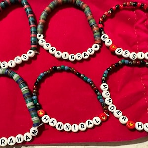 Könnte beinhalten: Fünf bunte Perlenarmbänder mit verschiedenen Wörtern, die darauf geschrieben sind. Die Wörter sind "Ravenstag", "Hannigram", "My Design", "Will Graham" und "Murder Husbands". Jedes Armband hat einen kleinen roten Herz-Anhänger.
