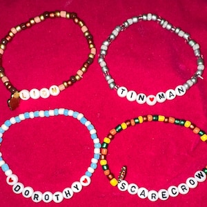 Può includere: Quattro braccialetti di perline con perline di diversi colori e perline di lettere bianche che compongono i nomi "Leone", "Uomo di latta", "Dorothy" e "Spaventapasseri".