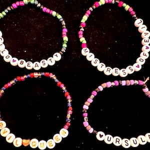 Könnte beinhalten: Vier Perlenarmbänder mit verschiedenen Perlenfarben und weißen Buchstabenperlen, die "Hello Beasty", "Mistress of Evil", "Evil Queen" und "Ursula" buchstabieren.