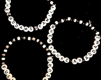 Op de afbeelding: Drie kralenarmbanden met zwarte en witte kralen die de zinnen "The Addams Family", "When You're An Addams" en "Full Disclosure" spellen.