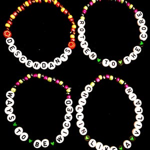 Op de afbeelding: Vier ronde kralenarmbanden met witte letterkralen en kleurrijke accentkralen. De armbanden tonen de teksten DESCENDANTS, ROTTEN TO THE CORE, WAYS TO BE WICKED en CHILLIN LIKE A VILLAIN, afgewisseld met kleine groene hartvormige kralen.