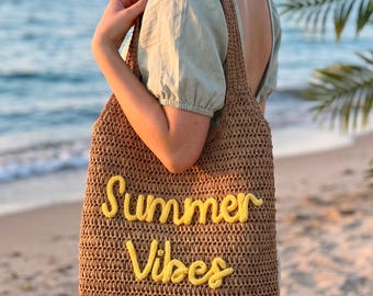 May include: A brown crocheted tote bag featuring the text Summer Vibes embroidered in yellow yarn on the front. The bag has a rectangular shape with a single wide shoulder strap and a textured, woven appearance made from natural-toned fiber.