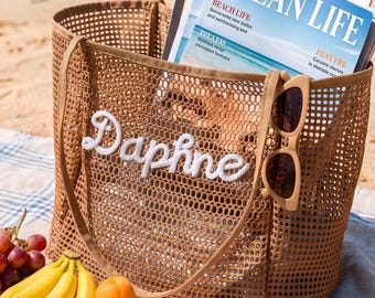 May include: A tan mesh tote bag featuring the name Daphne embroidered in white script on the front. The bag contains a magazine titled Ocean Life and has beige sunglasses hanging from one of the two shoulder straps, with fruit nearby.