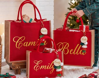 May include: Three red tote bags with natural brown side panels, each featuring gold cursive lettering reading Camila, Bella, or Emily. Each bag includes a round wooden personalized keychain with colorful tassels attached to the red fabric handles.