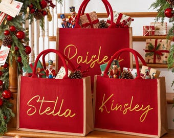 May include: Three red rectangular tote bags with tan trim and handles, each personalized with gold cursive text reading Paisley, Stella, and Kinsley. The bags are filled with small nutcracker figurines and wrapped gifts on a staircase.