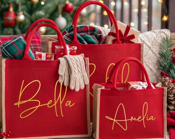 May include: Two red jute tote bags with sturdy handles, featuring the names Bella and Amelia in gold cursive script. The bags are filled with items including a pair of beige knit gloves, plaid fabric, and small wrapped gifts on a wooden surface.