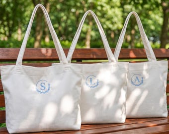 May include: Three off-white corduroy tote bags with two shoulder straps each. Each bag features a circular embroidered design in light blue thread, containing a single letter S, L, or A respectively, surrounded by a delicate leaf wreath motif.