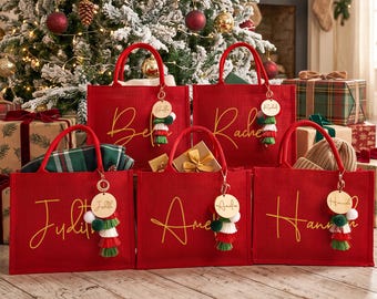 May include: Five rectangular red tote bags with two handles each, featuring gold cursive names printed on the front. Each bag includes a round wooden keychain tag with a name and a layered tassel in green, white, and red colors attached to the handle.