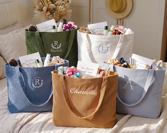 May include: Five corduroy tote bags in olive green, white, blue, mustard yellow, and light gray. Each bag features embroidered monograms or names, including Rachel, Charlotte, and Amelia, and is filled with assorted personal care items and cards.