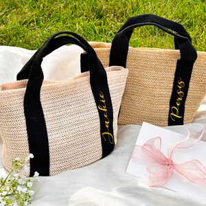 K&ouml;nnte beinhalten: Zwei gewebte Stroh-Tragetaschen mit schwarzen Griffen und B&auml;ndern. Eine Tasche tr&auml;gt den Namen "Jackie" in goldener Schrift, die andere "Persis". Die Taschen liegen auf einem wei&szlig;en Tuch mit gr&uuml;nem Gras im Hintergrund.