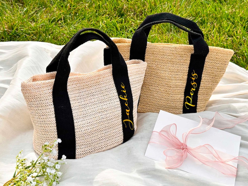 K&ouml;nnte beinhalten: Zwei gewebte Stroh-Tragetaschen mit schwarzen Griffen und B&auml;ndern. Eine Tasche tr&auml;gt den Namen "Jackie" in goldener Schrift, die andere "Persis". Die Taschen liegen auf einem wei&szlig;en Tuch mit gr&uuml;nem Gras im Hintergrund.