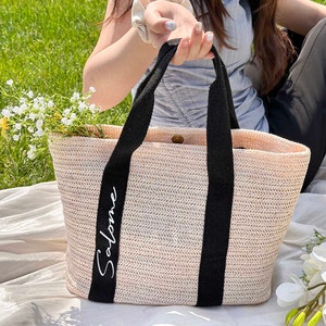 K&ouml;nnte beinhalten: Eine gewebte Stroh-Tragetasche mit schwarzen Griffen und dem Namen "Salome" in wei&szlig;er Schrift. Die Tasche ist mit wei&szlig;en Blumen gef&uuml;llt und liegt auf einer wei&szlig;en Decke auf einer grasbewachsenen Oberfl&auml;che. Die Tasche hat eine hellbeige Farbe.