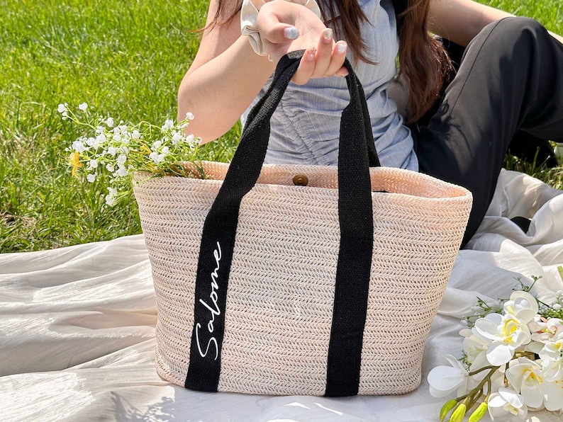 K&ouml;nnte beinhalten: Eine gewebte Stroh-Tragetasche mit schwarzen Griffen und dem Namen "Salome" in wei&szlig;er Schrift. Die Tasche ist mit wei&szlig;en Blumen gef&uuml;llt und liegt auf einer wei&szlig;en Decke auf einer grasbewachsenen Oberfl&auml;che. Die Tasche hat eine hellbeige Farbe.