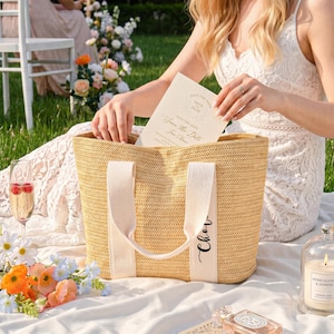 K&ouml;nnte beinhalten: Eine Stroh-Tragetasche mit cremefarbenen Griffen und dem Namen "Cher" in Schreibschrift. Die Tasche liegt auf einer wei&szlig;en Decke mit Blumen, Sektgl&auml;sern und einer Kerze. Eine "Save the Date"-Karte wird gehalten.