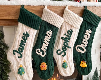 May include: Four cable-knit Christmas stockings hanging in a row, alternating between cream and dark green colors. Each stocking features a folded cuff, a personalized name in cursive script, and a small decorative felt applique at the bottom.