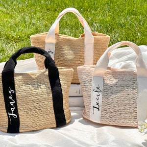 K&ouml;nnte beinhalten: Drei gewebte Stroh-Tragetaschen mit personalisierten Namen auf den Tr&auml;gern. Eine Tasche hat schwarze Tr&auml;ger und den Namen "Janey" in wei&szlig;er Schrift. Die anderen beiden Taschen haben cremefarbene Tr&auml;ger mit dem Namen "Lucile" in wei&szlig;er Schrift.