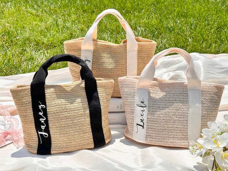 K&ouml;nnte beinhalten: Drei gewebte Stroh-Tragetaschen mit personalisierten Namen auf den Tr&auml;gern. Eine Tasche hat schwarze Tr&auml;ger und den Namen "Janey" in wei&szlig;er Schrift. Die anderen beiden Taschen haben cremefarbene Tr&auml;ger mit dem Namen "Lucile" in wei&szlig;er Schrift.